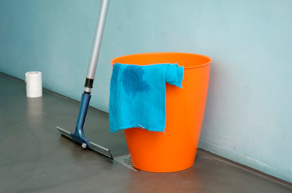 A close-up of an orange plastic bucket placed on a smooth, grey floor near a light blue wall, containing a blue microfiber cleaning cloth draped over the edge. A mop with a silver handle and blue head is leaning against the bucket, positioned in front of a small white roll of toilet paper or paper towels on the floor. The scene is well-lit, emphasizing the cleanliness of the floor and surfaces, typical of professional surface cleaning or deep cleaning in a residential or commercial setting. The environment showcases tools used by Cleaners N4 for effective sanitisation and maintenance of tiled or vinyl flooring.