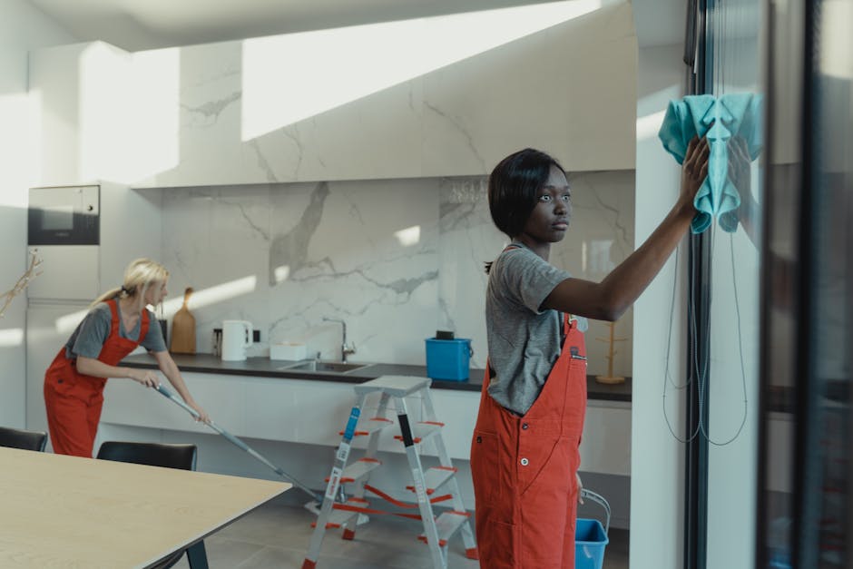 Two professional cleaners in a modern kitchen, both wearing red aprons and grey t-shirts. The cleaner in the foreground, a woman with short black hair, is wiping a glass surface with a blue microfiber cloth, ensuring streak-free cleanliness. The second cleaner, a woman with blonde hair, is mopping the tiled floor using a mop, with a white bucket nearby. The kitchen features a white marble backsplash, stainless steel appliances, and a wooden table, all under bright, natural lighting. The surfaces are spotless and shiny, indicating thorough surface cleaning and sanitisation. Modern cleaning tools and materials, including a ladder and spray bottles, are visible, emphasizing the professional cleaning process. This image exemplifies detailed domestic cleaning, highlighting the importance of surface and floor hygiene, as promoted by Cleaners N4 for local Finsbury Park residents seeking reliable, transparent cleaning services without hidden charges.