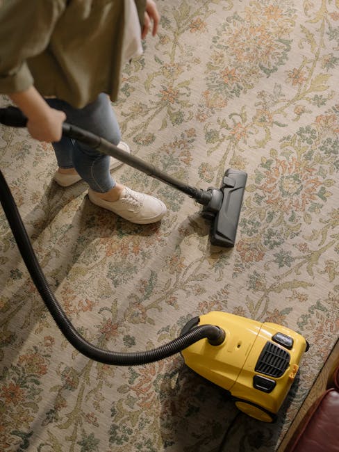 A person is performing surface cleaning on a patterned area rug using a vacuum cleaner with a long hose attachment. The individual is dressed in casual clothing, including beige shoes, and is focused on thoroughly cleaning the rug, which features a floral design with shades of beige, green, and orange. The scene takes place in a well-lit room, highlighting the clean and maintained condition of the carpet. The vacuum cleaner, a yellow model, is connected to a power source, and the person's hand guides the hose attachment across the rug’s surface. This image illustrates professional deep cleaning and sanitisation processes conducted by Cleaners N4 as part of their domestic cleaning and carpet care services, emphasizing meticulous maintenance of residential flooring surfaces.