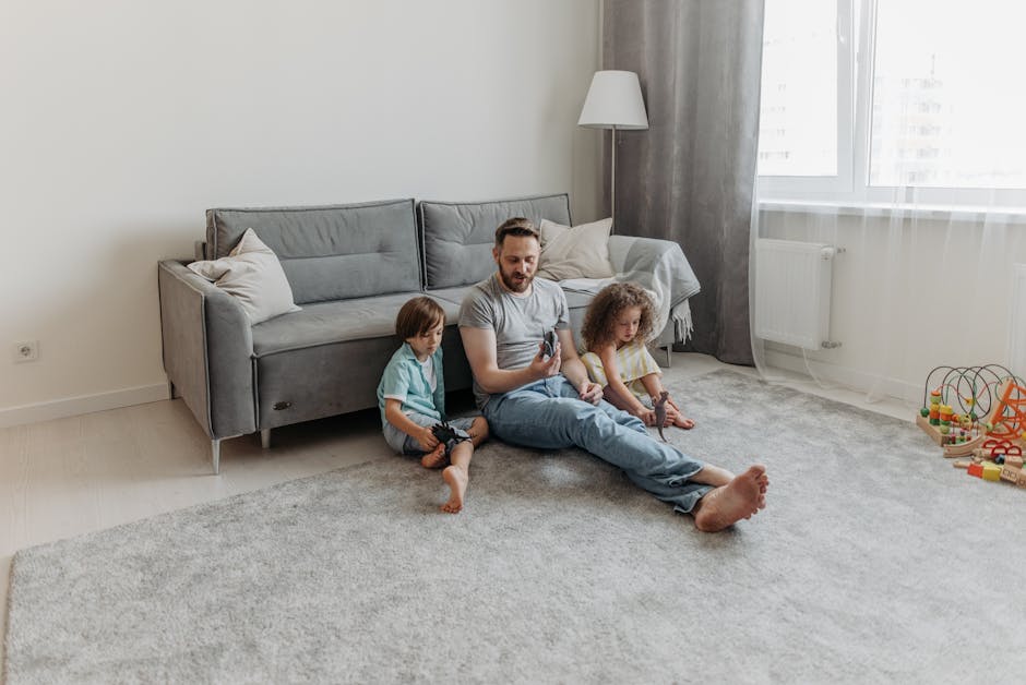 A father seated on a light grey, plush carpet in a modern living room with his two young children, a boy and a girl, sitting beside him. The father is holding a smartphone, while the children hold small toys. Behind them is a grey fabric sofa with matching cushions, positioned against a white wall. To the right, a tall standing lamp with a white lampshade stands next to a large window with sheer white curtains, allowing natural light to fill the room. The carpet appears clean and well-maintained, reflecting the expertise of Cleaners N4 in surface cleaning and deep cleaning services for domestic spaces. Visible toys on the floor and the bright, tidy environment emphasize the importance of hygiene and regular cleaning in creating a safe and comfortable home. The room's neutral color palette, comprising shades of grey and white, enhances the overall sense of cleanliness and order.