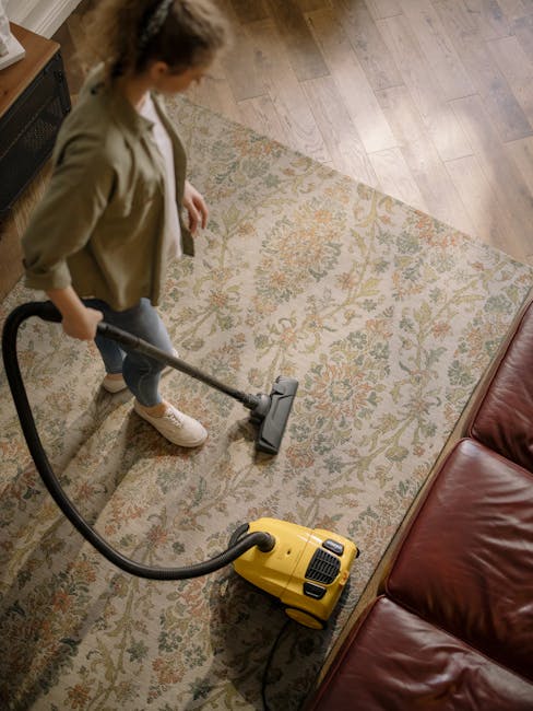 A person is performing surface cleaning on a patterned area rug using a vacuum cleaner with a long hose attachment. The individual is dressed in casual clothing, including beige shoes, and is focused on thoroughly cleaning the rug, which features a floral design with shades of beige, green, and orange. The scene takes place in a well-lit room, highlighting the clean and maintained condition of the carpet. The vacuum cleaner, a yellow model, is connected to a power source, and the person's hand guides the hose attachment across the rug’s surface. This image illustrates professional deep cleaning and sanitisation processes conducted by Cleaners N4 as part of their domestic cleaning and carpet care services, emphasizing meticulous maintenance of residential flooring surfaces.