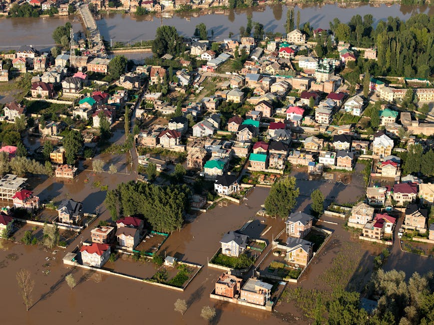 An aerial view of a residential neighborhood showing houses with various colored rooftops and garden areas, partially submerged in floodwaters following heavy rainfall. Streets are flooded, with water covering pathways and driveways, creating a muddy, brown appearance. Trees and greenery are visible along the streets and in backyards, contrasting with the flooded surfaces. The scene depicts the effects of flooding common in Finsbury Park, N4, highlighting the need for effective surface cleaning and sanitisation to restore hygiene and safety. Cleaners N4 specialises in emergency flood cleaning for homes affected by such common problems in the area, ensuring thorough cleaning and debris removal.