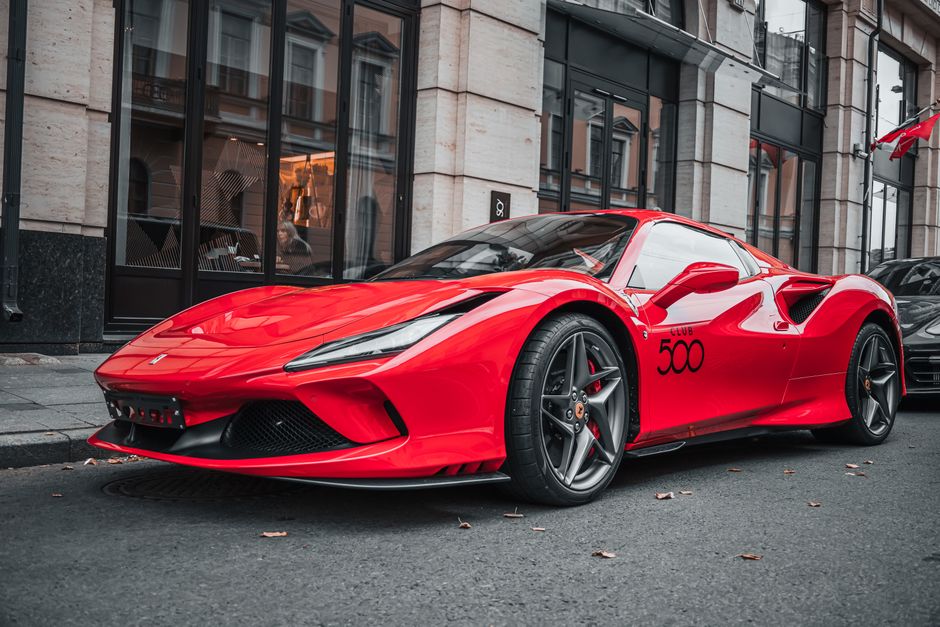 A bright red sports car with a sleek, aerodynamic design parked on a city street in front of a modern building with large glass windows. The vehicle features smooth, shiny surfaces, including the hood, doors, and side panels, which are clean and well-maintained, reflecting surrounding buildings and streetlights. The black alloy wheels are detailed and free of dirt or debris. The car's windows are clear, and the bodywork displays the number '500' in black on the side. The street appears tidy with minimal fallen leaves, and a subtle urban backdrop is visible, emphasizing the vehicle's polished appearance. This image showcases a luxury vehicle, likely after thorough surface cleaning and detailing, aligning with the concept of high-quality automotive or surface cleaning services offered by Cleaners N4, a specialist in domestic and commercial cleaning.