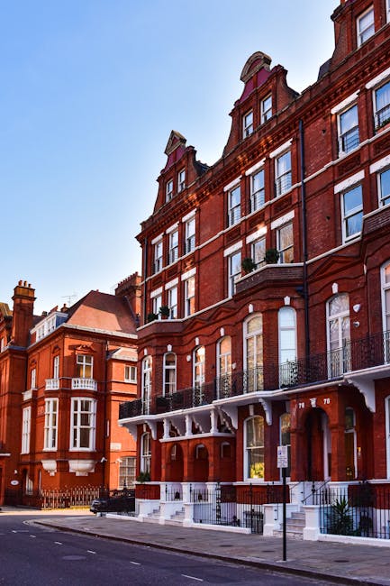 An exterior view of a historic red brick residential building with multiple floors featuring large sash windows and decorative architectural details. The ground floor has white-painted timber accents supporting a small balcony with ornate black wrought iron railings. The windows on the upper floors are tall, with some adorned with small balconies and potted plants. The building is situated on a quiet street, with a clear blue sky overhead and neighboring similar structures visible in the background. This image emphasizes the classic Victorian-style architecture, clean and well-maintained surfaces, and the sunny weather, reflecting the type of environment where professional domestic cleaning services by Cleaners N4 can support maintaining such beautiful homes.