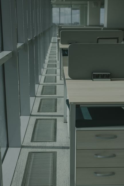 A row of white office desks with attached grey dividers, positioned along a windowed wall in a modern workspace. The desks have smooth, laminated surfaces with some visible electrical outlets. Beneath the desks, there are grey and black cable management trays. The floor is covered with light grey carpet, and there are floor vents with metal grilles aligned parallel to the desks, suggesting surface cleaning and ventilation maintenance. Natural light filters through the large windows, illuminating the space evenly. The overall appearance is clean, tidy, and well-maintained, reflecting professional cleaning standards in a commercial office environment, as provided by Cleaners N4.