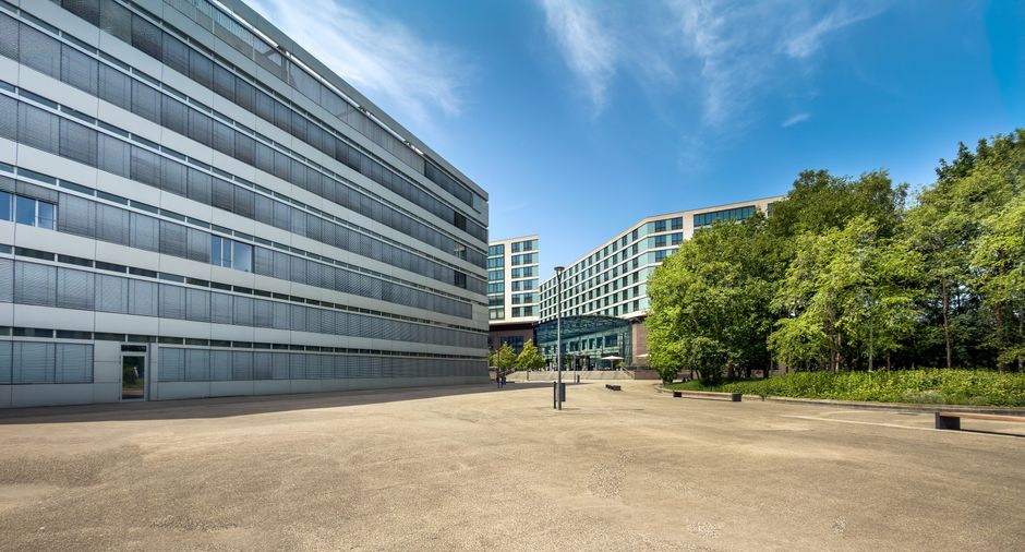 An exterior view of a modern office complex featuring large glass windows and sleek metal cladding on multi-storey buildings, with an expansive paved courtyard in the foreground. To the right, there are lush green trees and well-maintained garden beds, providing a contrast to the contemporary architecture. The scene is illuminated by bright, natural sunlight under a clear blue sky, highlighting the clean and orderly appearance of the outdoor space. The area appears tidy and well-kept, with no visible debris or dirt on the paving or building facades, reflecting professional cleaning and maintenance services. The image exemplifies the type of environment that Cleaners N4 specialises in maintaining, emphasizing surface cleanliness and overall hygiene for commercial properties in the Finsbury Park area.