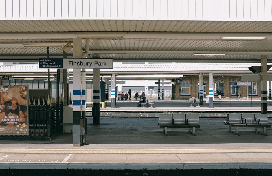 A view of Finsbury Park train station platform showing clean, tiled surfaces with polished metal benches and blue-and-white striped pillars supporting a beige, corrugated roof. The platform appears well-maintained, free of dust or litter, and is illuminated by natural daylight. In the background, visible signs indicate directions and station amenities, with a few passengers seated on benches or walking along the platform. The overall scene emphasizes the clean, hygienic environment typical of professional station maintenance, reflecting the quality service offered by Cleaners N4 for domestic and commercial cleaning, including surface cleaning and sanitisation tasks.