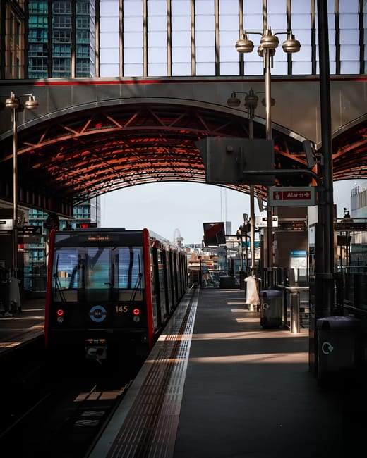 Photograph of Finsbury Park Station platform with a modern train arriving or departing, featuring sleek black and red exterior. The platform surface is clean with tactile paving strips along the edge for safety. Adjacent to the train, there are benches, a trash bin, and signage including an 'Alarm' indicator. The station's covered area has a contemporary design with a curved roof made of glass and metal, illuminated by hanging pendant lights. Surrounding the station, tall glass buildings reflect the sky and urban environment, creating a bright, well-maintained appearance. This scene exemplifies professional cleaning standards for public transportation hubs, highlighting surfaces such as the platform, glass, and metallic fixtures, emphasizing cleanliness and maintenance typical of service providers like Cleaners N4, who specialise in sanitisation and deep cleaning for commercial spaces.