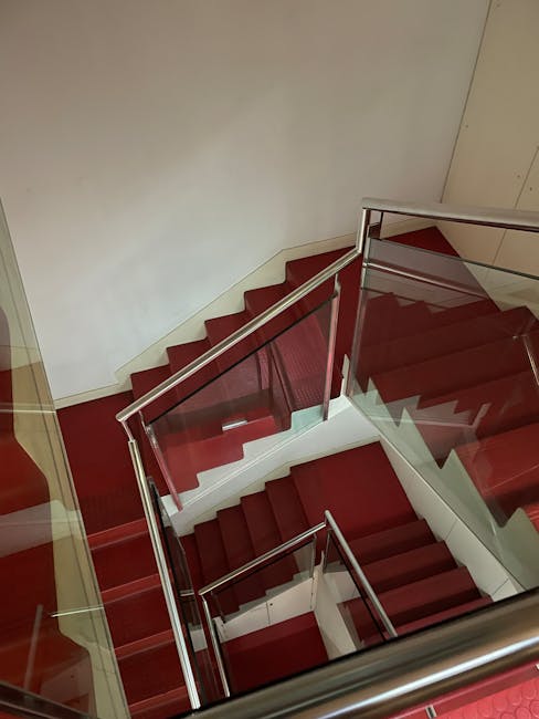 Aerial view of a modern staircase with red carpeted steps, metal handrails, and glass panels enclosing the sides. The stairs are illuminated by overhead lighting, and the walls adjacent are painted wh