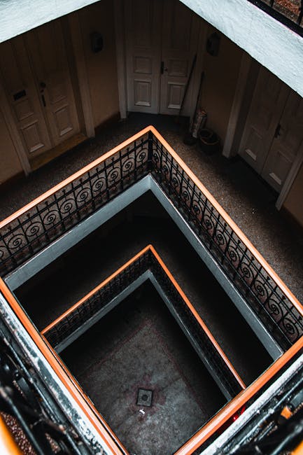 Aerial view of a modern staircase with red carpeted steps, metal handrails, and glass panels enclosing the sides. The stairs are illuminated by overhead lighting, and the walls adjacent are painted wh